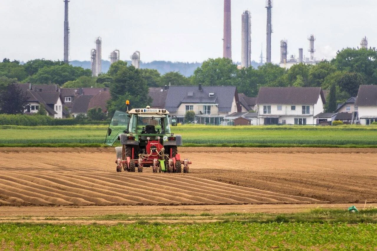 Tractor verde ara surcos en un campo cultivado con viviendas y chimeneas industriales al fondo.