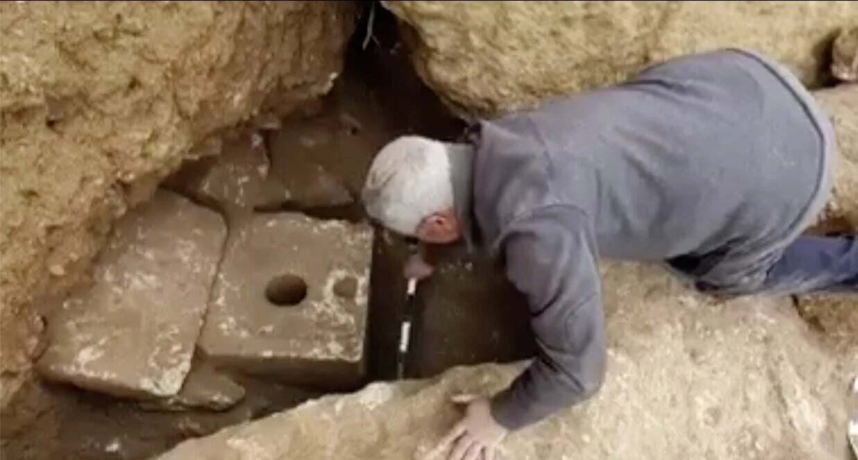 Un arqueólogo se inclina para inspeccionar de cerca una letrina de piedra tallada descubierta en una excavación arqueológica.