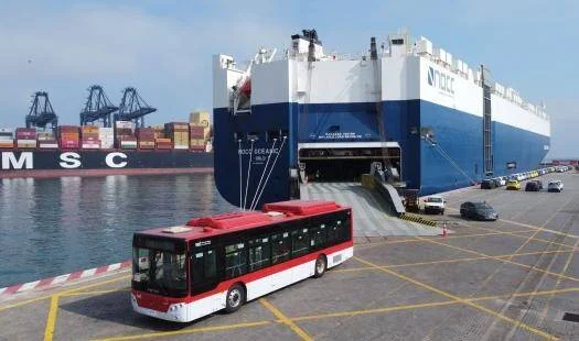Un autobús urbano rojo y blanco desembarca de un buque de carga ro-ro en un puerto marítimo.