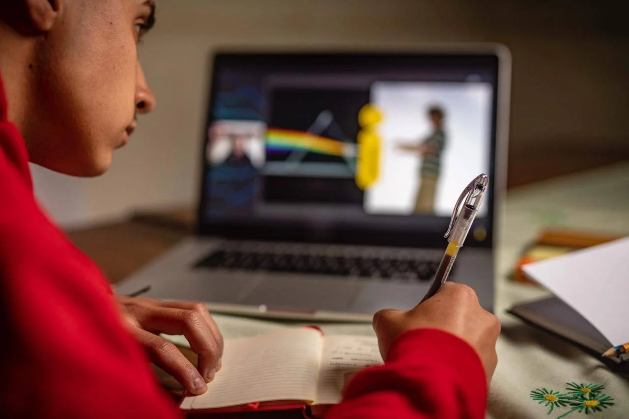 Joven con buzo rojo toma apuntes en un cuaderno mientras sigue una clase de física en su laptop.