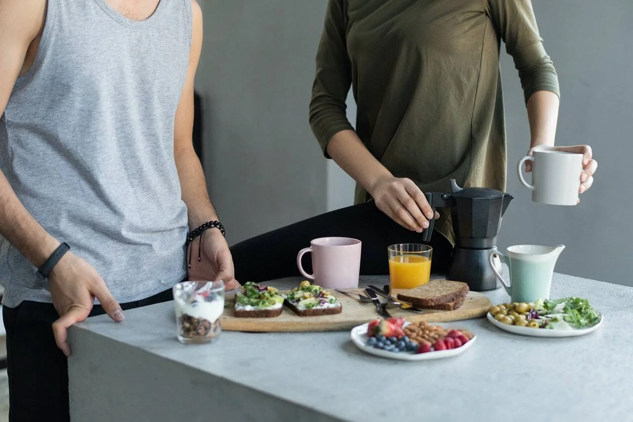 Dos personas de pie ante una mesa con desayuno saludable: tostadas con verduras, frutas, jugo de naranja y café recién preparado.