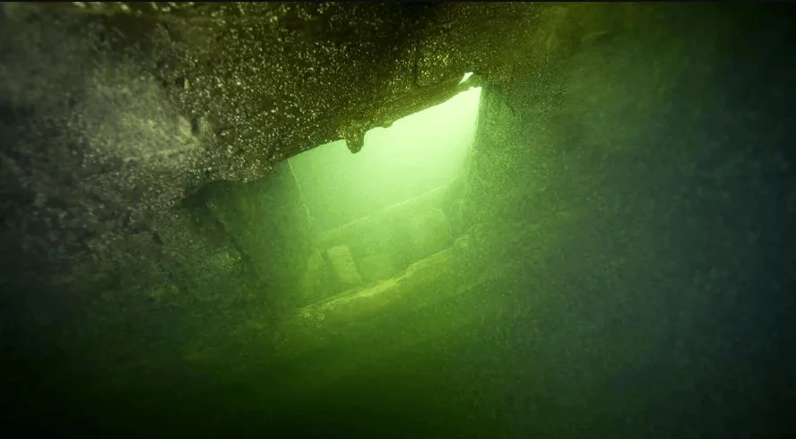 Vista submarina de una abertura en una estructura rocosa o metálica con luz verde filtrándose desde el interior.