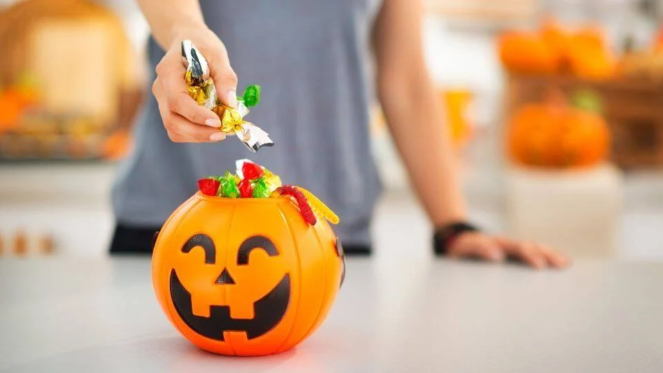 Una mano toma caramelos coloridos de un cubo con forma de calabaza de Halloween sobre una mesa.