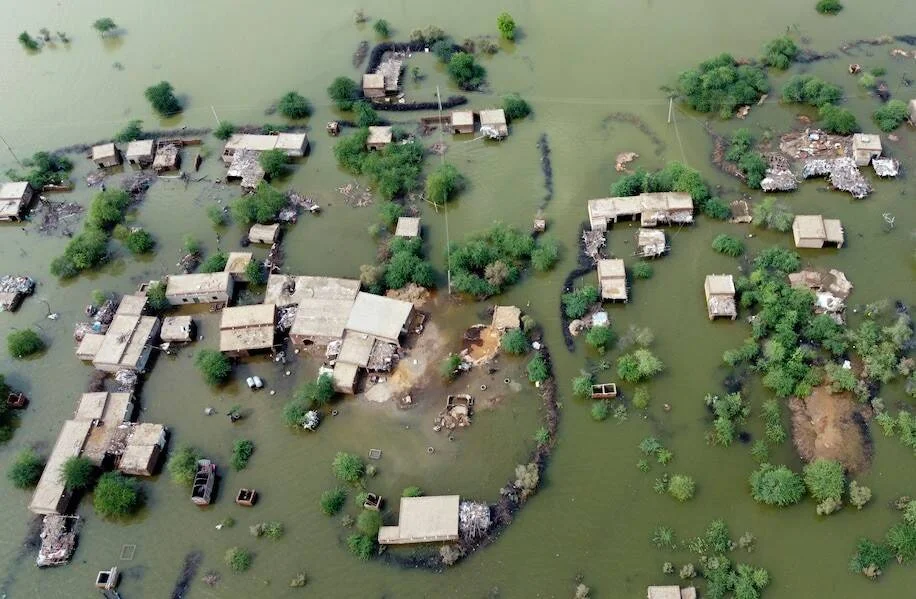 Fotografía aérea de un poblado rural con viviendas y árboles rodeados por aguas de inundación de color verdoso.