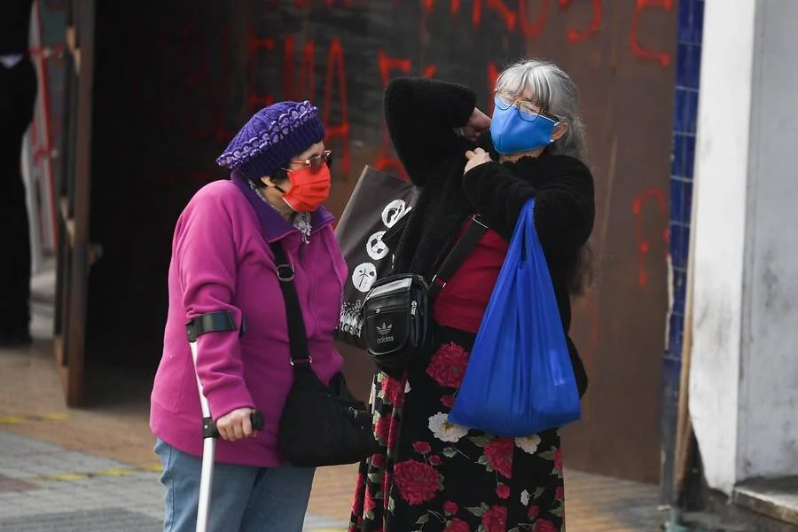 Tres mujeres con barbijos conversan en la vereda, una de ellas apoya en un bastón.