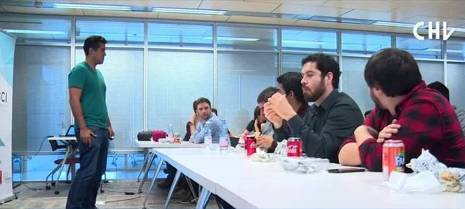 Un joven de pie expone ante un grupo de personas sentadas en una mesa de reuniones con bebidas sobre la mesa.