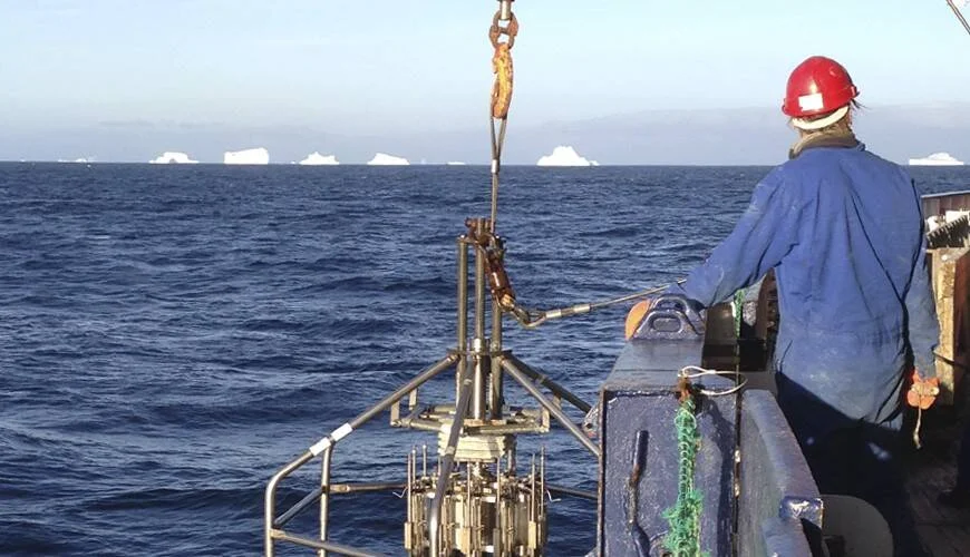 Investigador con casco rojo manipula instrumental científico suspendido desde un barco en aguas polares con icebergs al fondo.