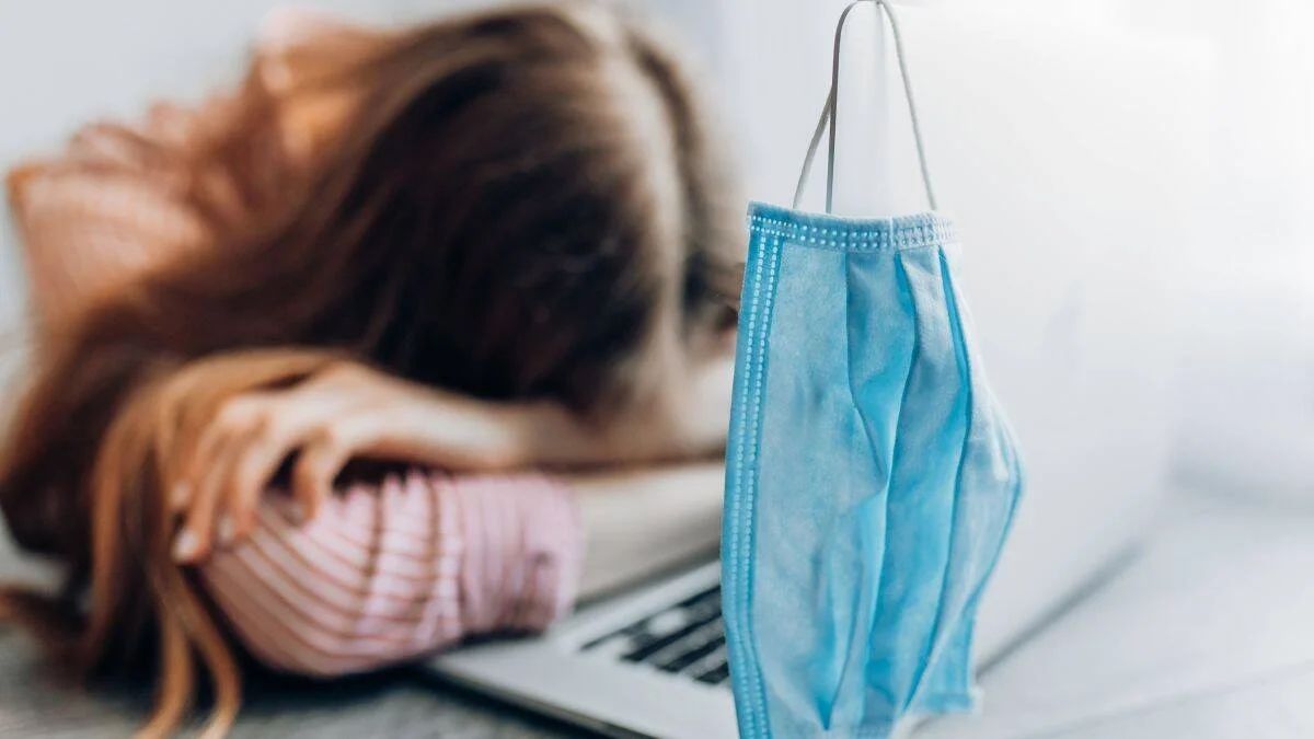 Mujer recostada sobre un escritorio con laptop, con un barbijo quirúrgico azul en primer plano, evocando cansancio por enfermedad.