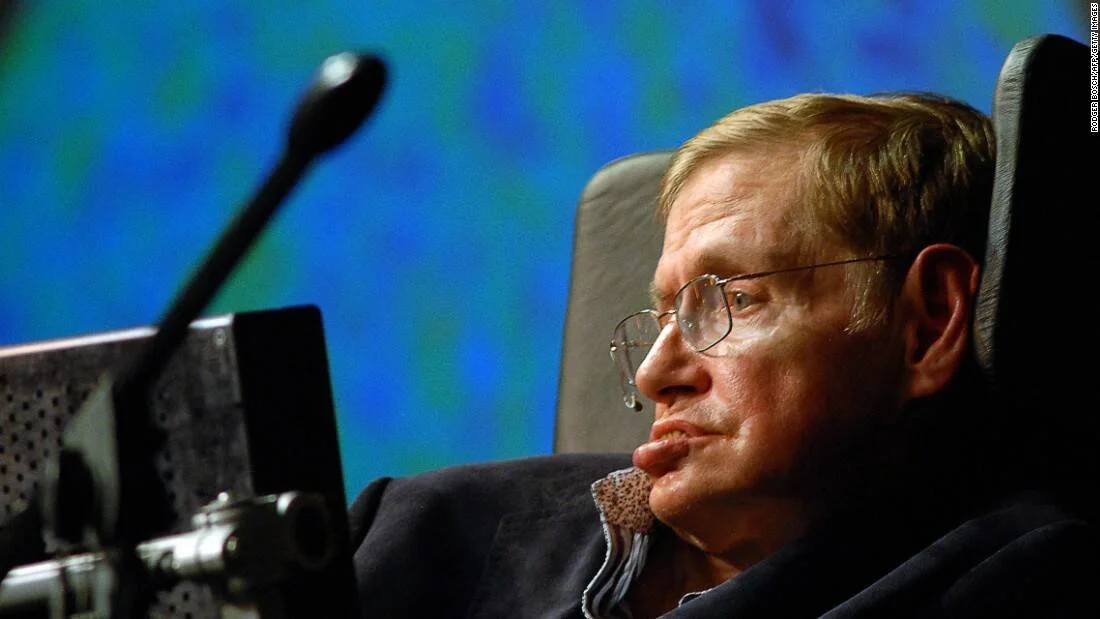 Stephen Hawking sentado en su silla motorizada frente a un micrófono, con fondo iluminado en azul y verde.