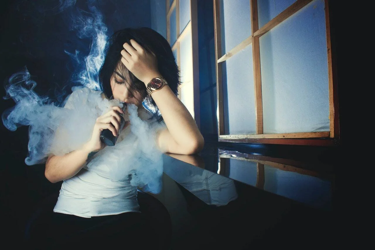 Mujer joven usando un cigarrillo electrónico junto a una ventana, rodeada de vapor, con gesto de estrés.