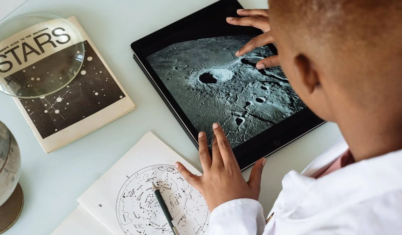 Un niño señala la superficie lunar en una tablet, junto a un mapa de constelaciones y un libro sobre estrellas.