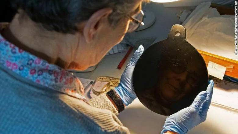 Investigador con guantes examina de cerca un espejo negro antiguo de forma ovalada bajo luz artificial en un laboratorio.