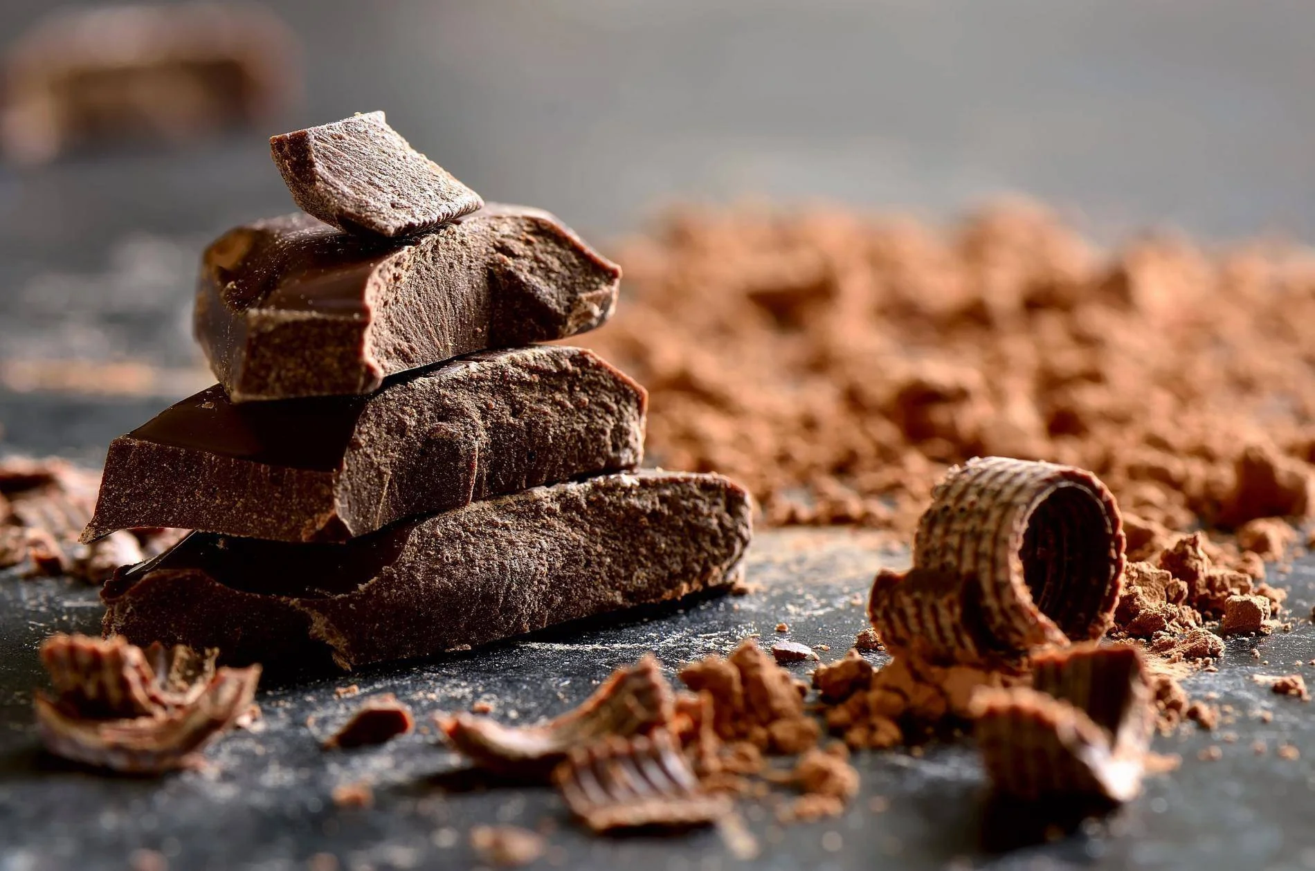 Piezas de chocolate negro apiladas rodeadas de virutas y polvo de cacao sobre superficie oscura.