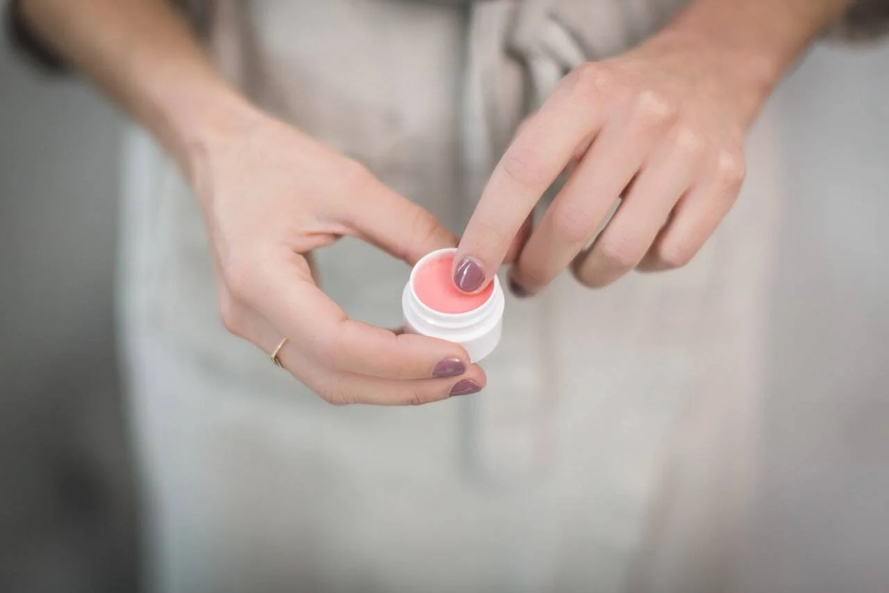 Una persona toma con el dedo un bálsamo labial de color rosado contenido en un pequeño tarro blanco.