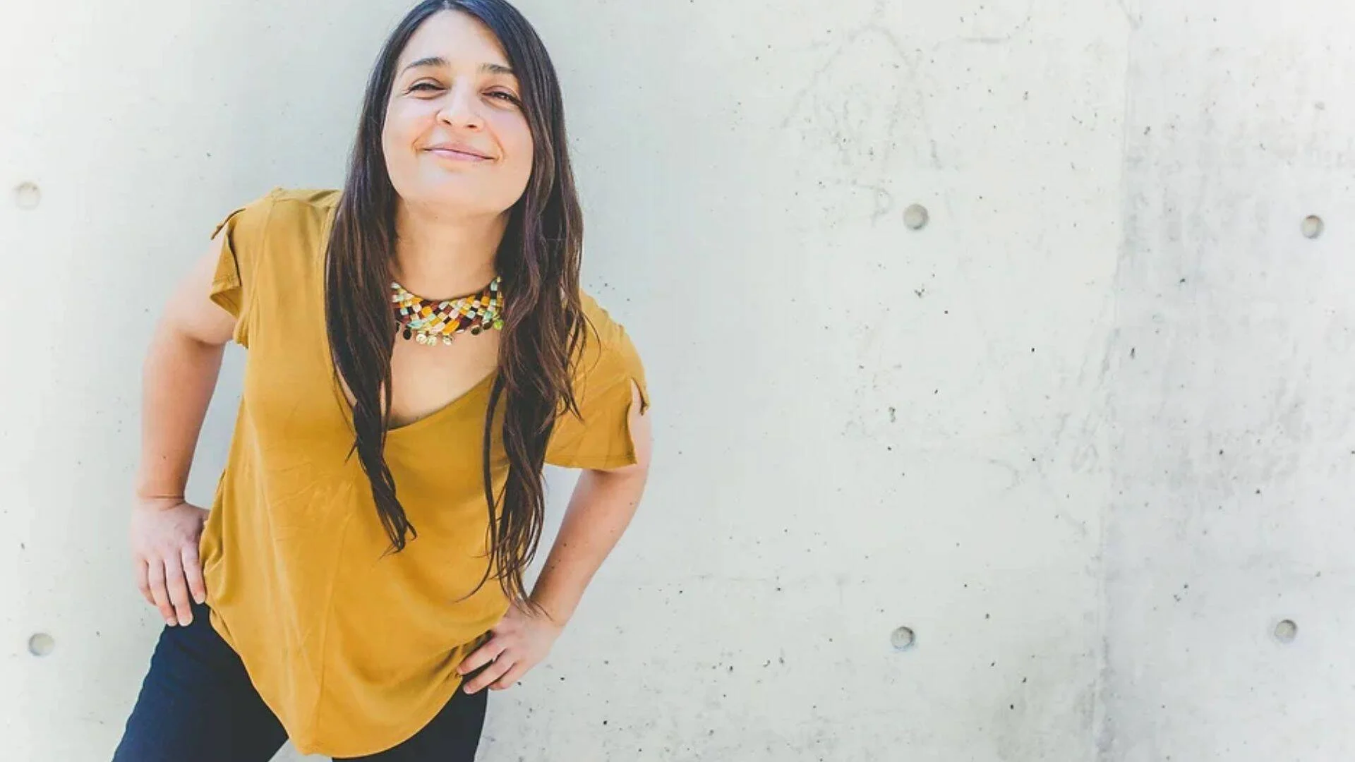 Mujer de cabello largo oscuro, blusa amarilla mostaza y collar artesanal, sonriendo ante una pared de hormigón.