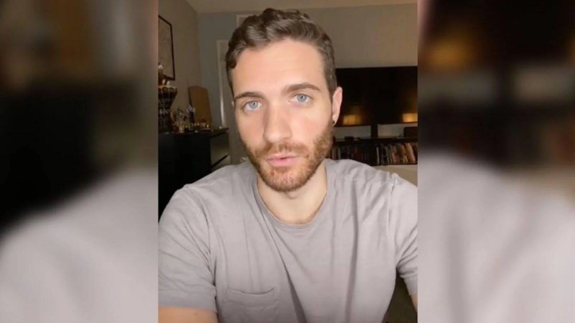 Hombre joven con barba corta y ojos azules graba un video desde su casa, con estanterías al fondo.