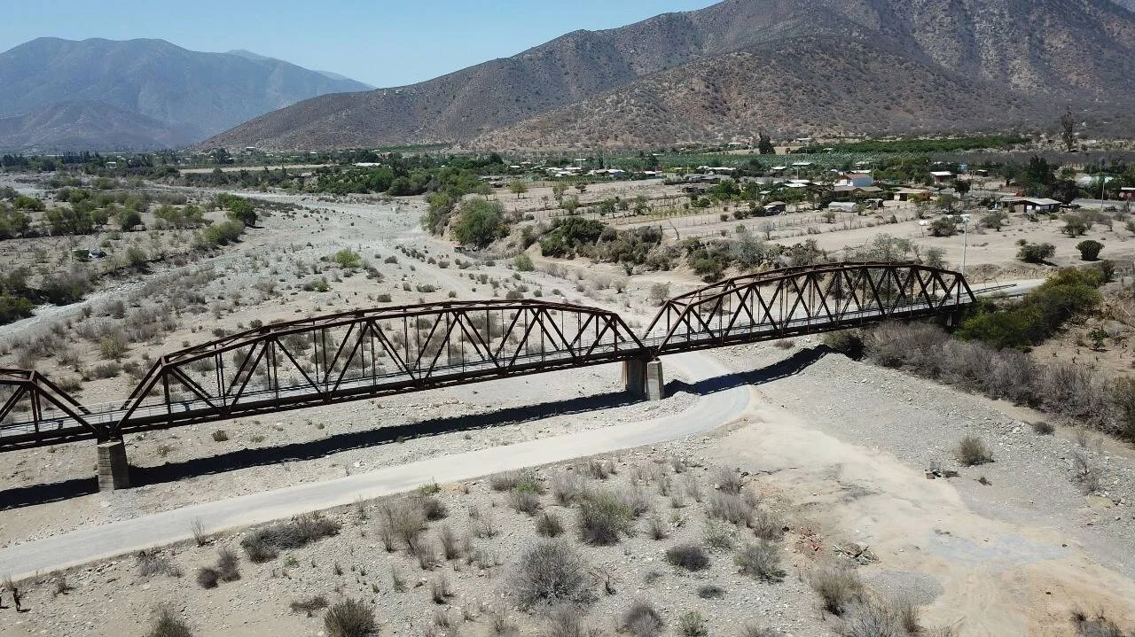 Puente de estructura metálica tipo cercha cruza un cauce de río completamente seco, rodeado de terreno árido y montañas al fondo.