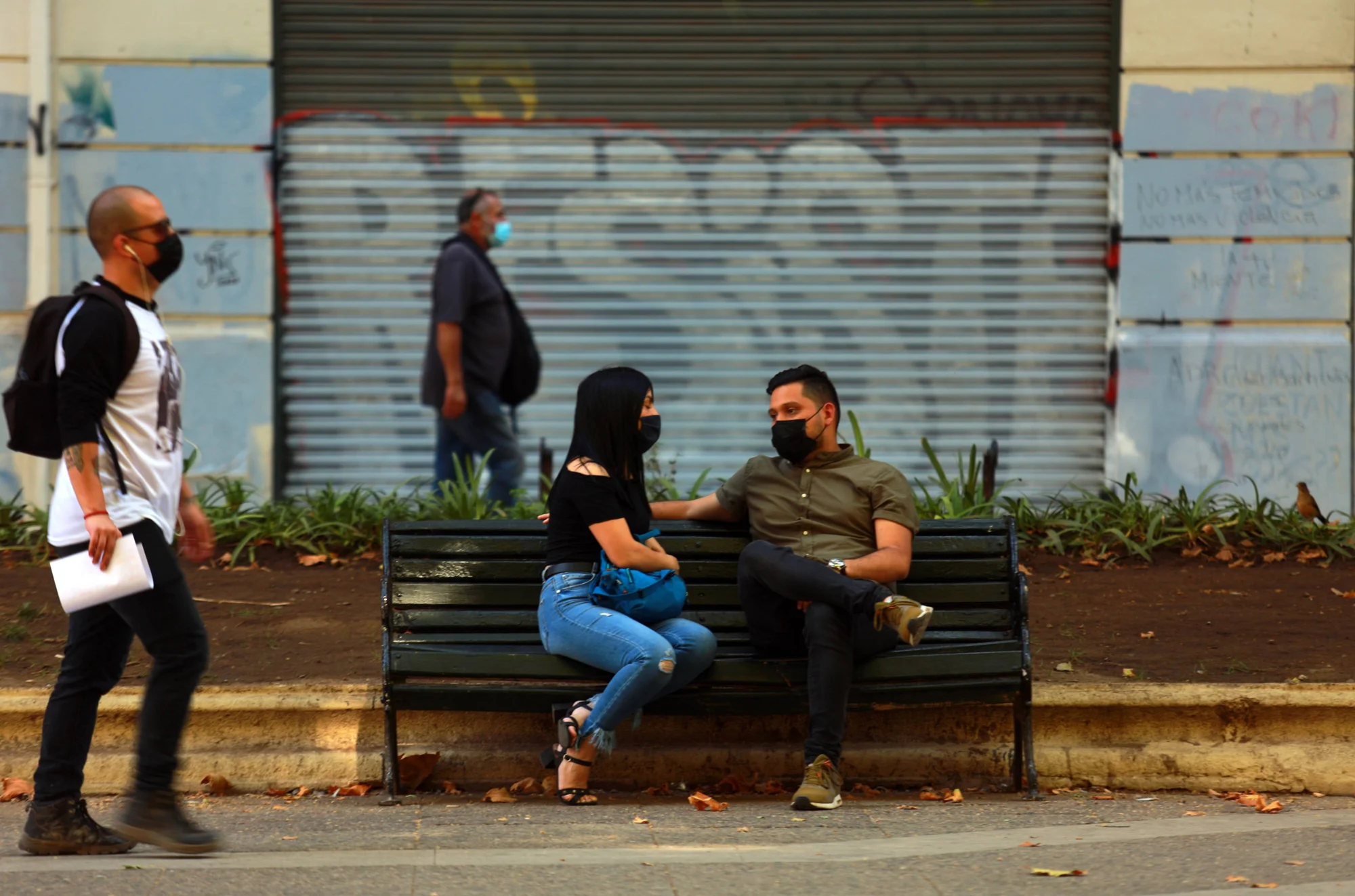 Una pareja con barbijos conversa sentada en un banco de plaza, mientras transeúntes con mascarilla pasan por la vereda.