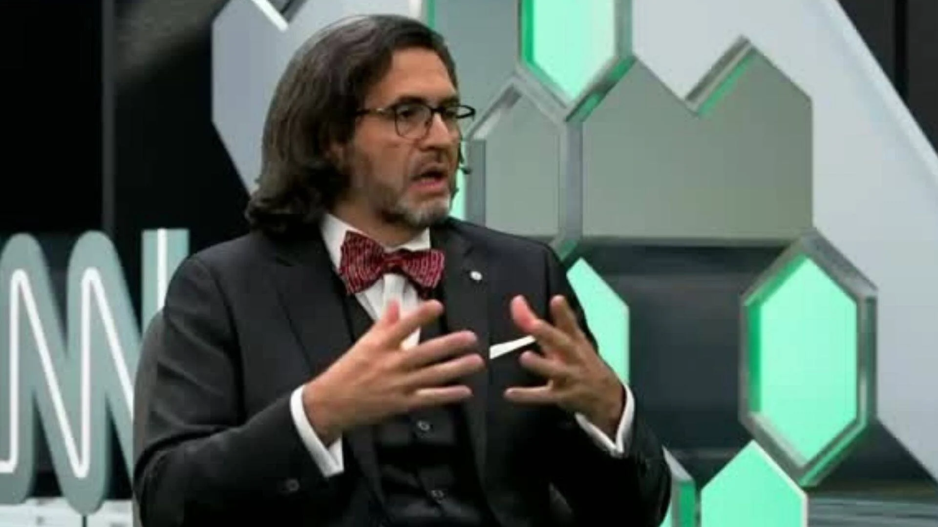 Hombre con cabello largo, gafas y moño rojo gesticula mientras habla en un estudio de televisión con fondo hexagonal verde.