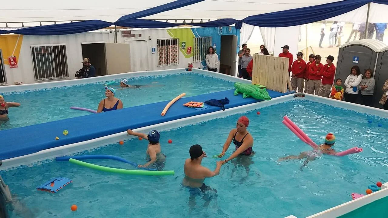 Instructores y niños practican natación con flotadores de colores en una piscina portátil, mientras autoridades observan desde afuera.