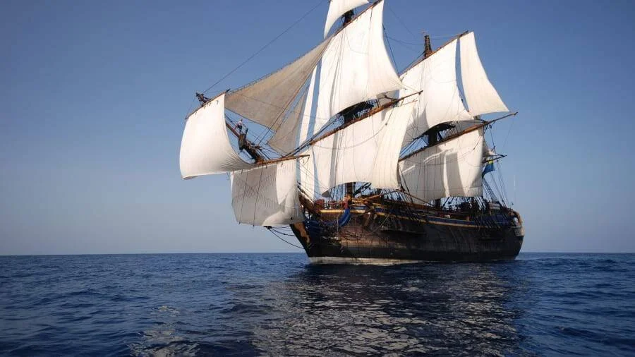 Gran velero de época con velas blancas desplegadas navega en un mar tranquilo bajo cielo despejado.