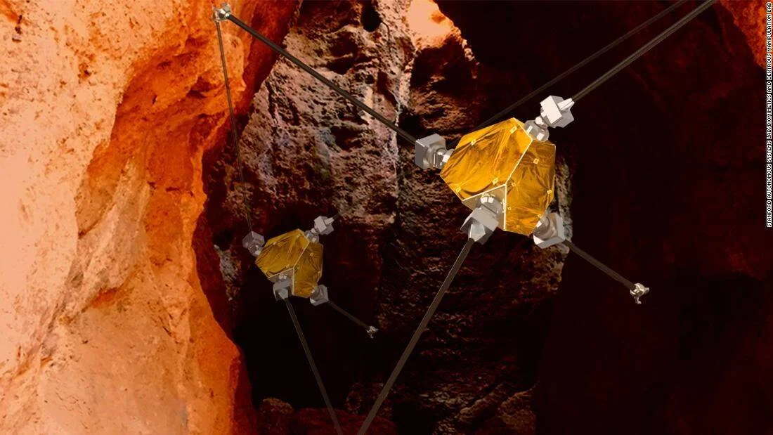 Concepto artístico de dos robots suspendidos por cables explorando una cueva de roca rojiza en Marte.