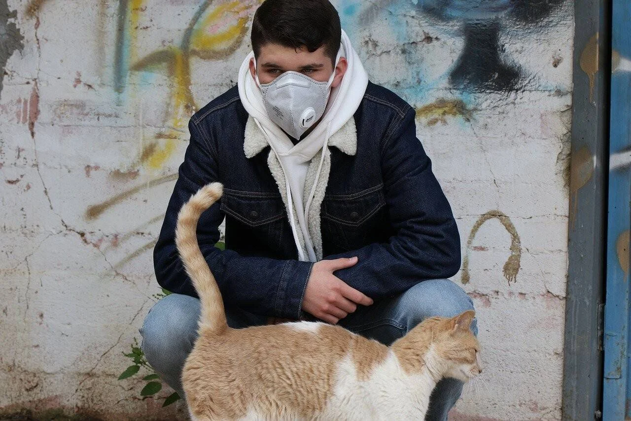 Un joven con mascarilla respiratoria y campera de jean acaricia un gato naranja y blanco en la calle.