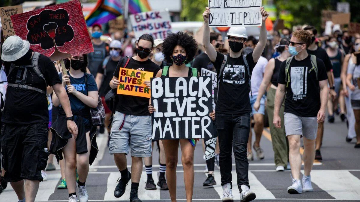 Manifestantes con mascarillas marchan sosteniendo carteles que dicen 'Black Lives Matter' y 'Defund the Police' en una protesta callejera.