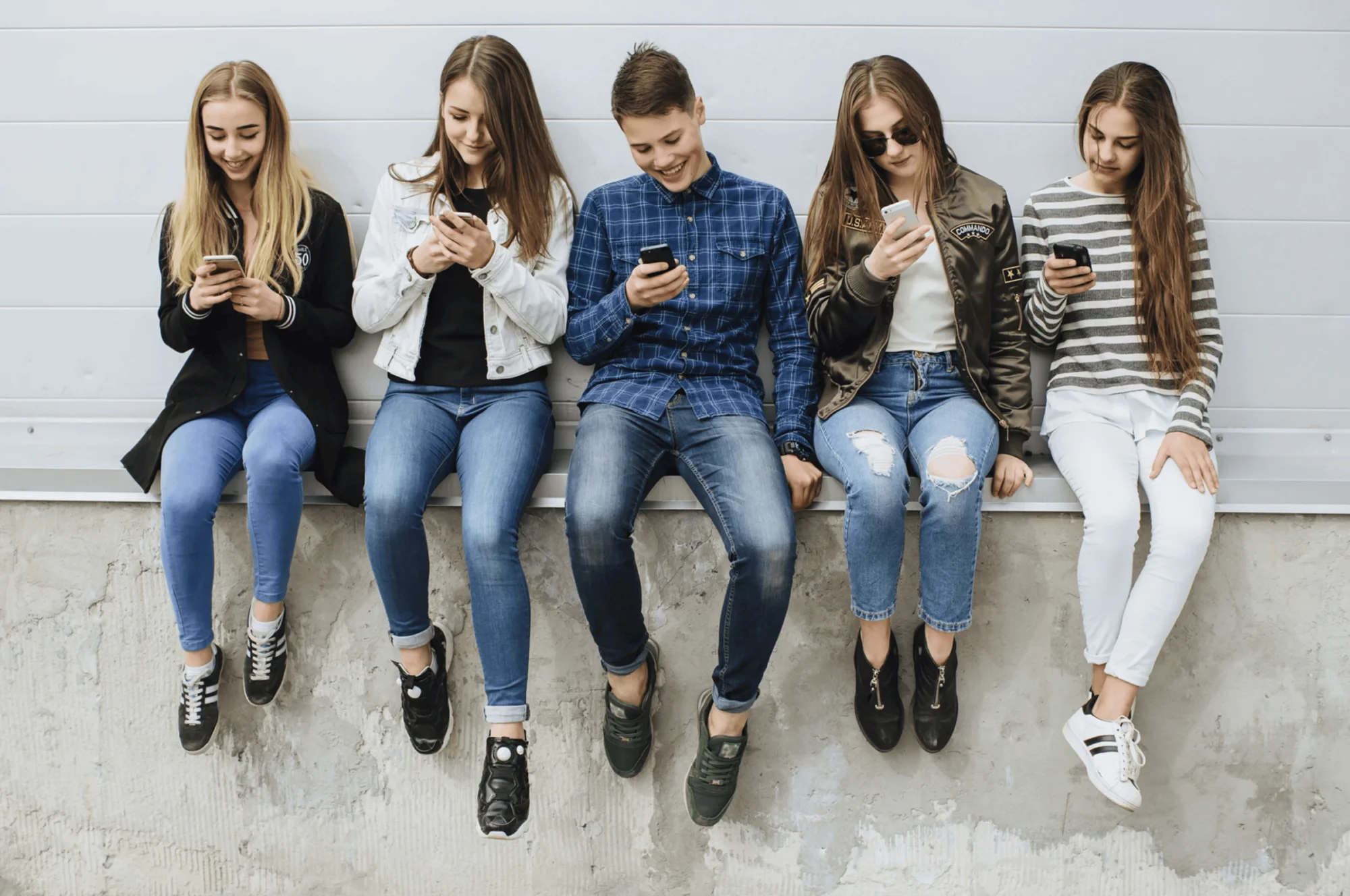 Cinco adolescentes sentados en fila sobre un muro, cada uno mirando su teléfono móvil.