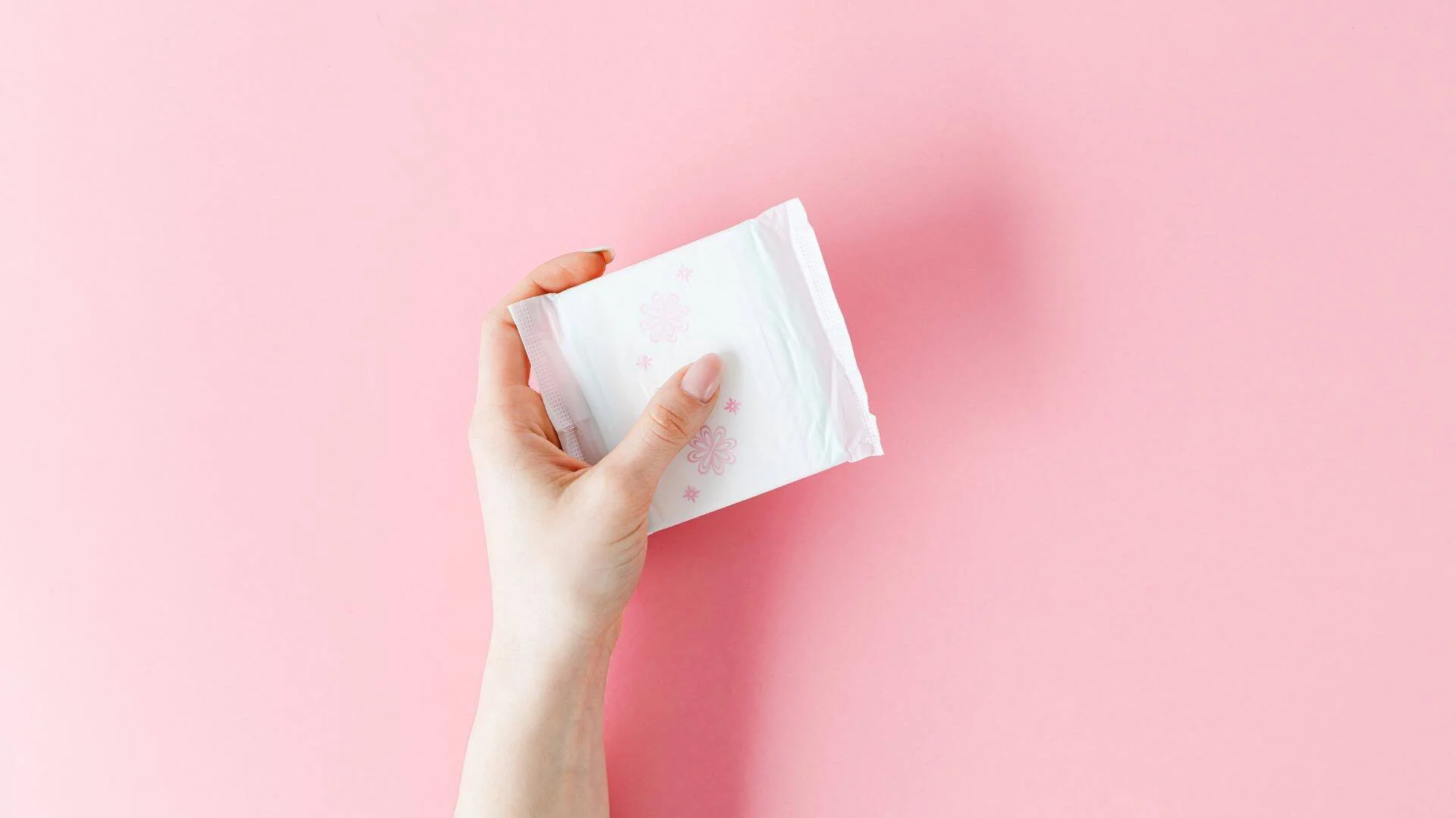 Una mano sostiene una compresa menstrual en su envoltorio blanco con motivos florales rosas, sobre fondo rosa.