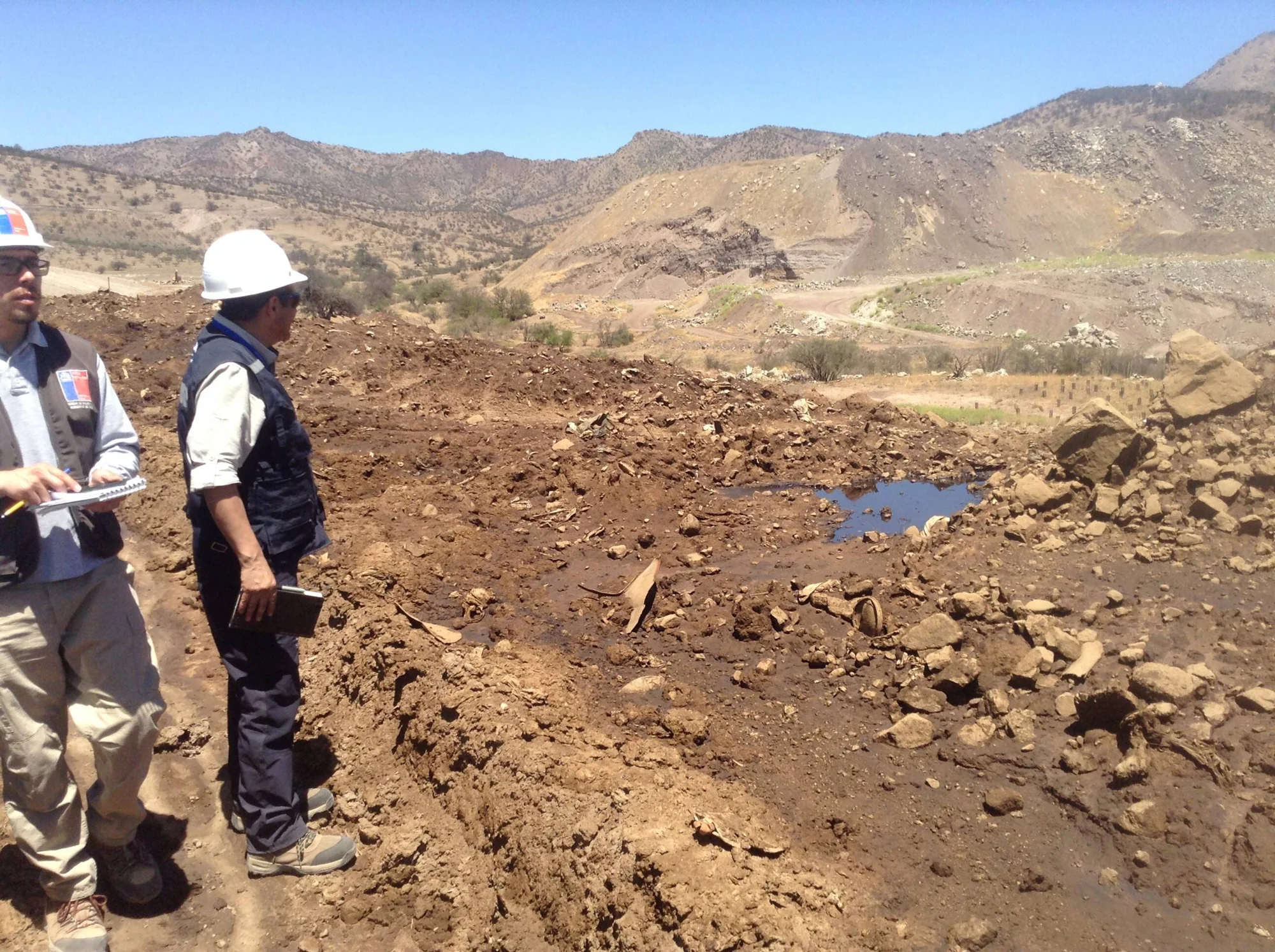 Dos técnicos con cascos de seguridad inspeccionan un terreno removido y lodoso en zona minera árida con cerros al fondo.