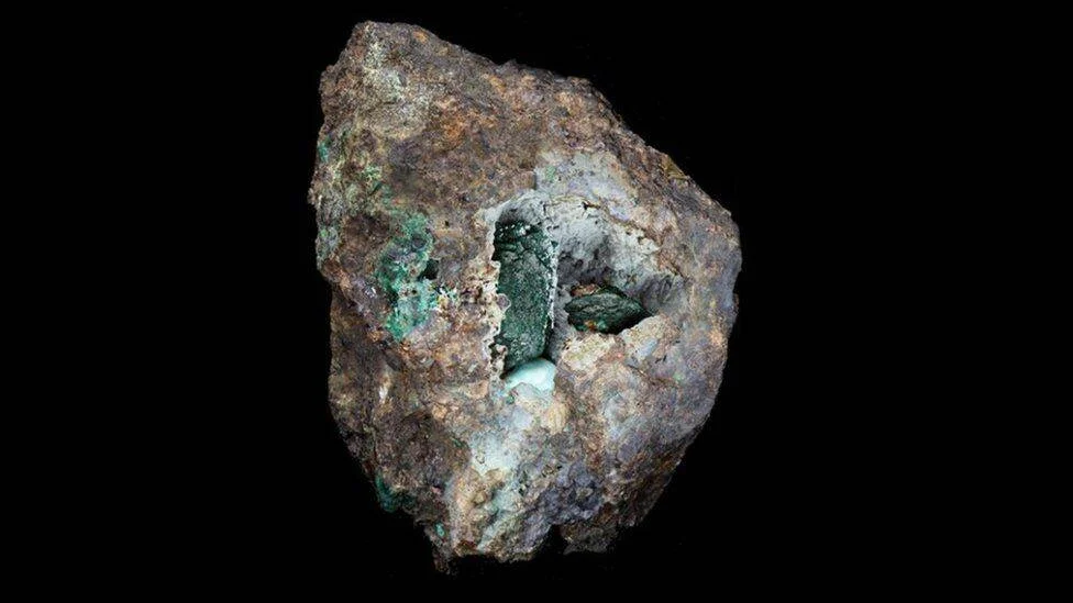 Fragmento de roca con minerales de color verde, posiblemente malaquita o cobre oxidado, sobre fondo negro.
