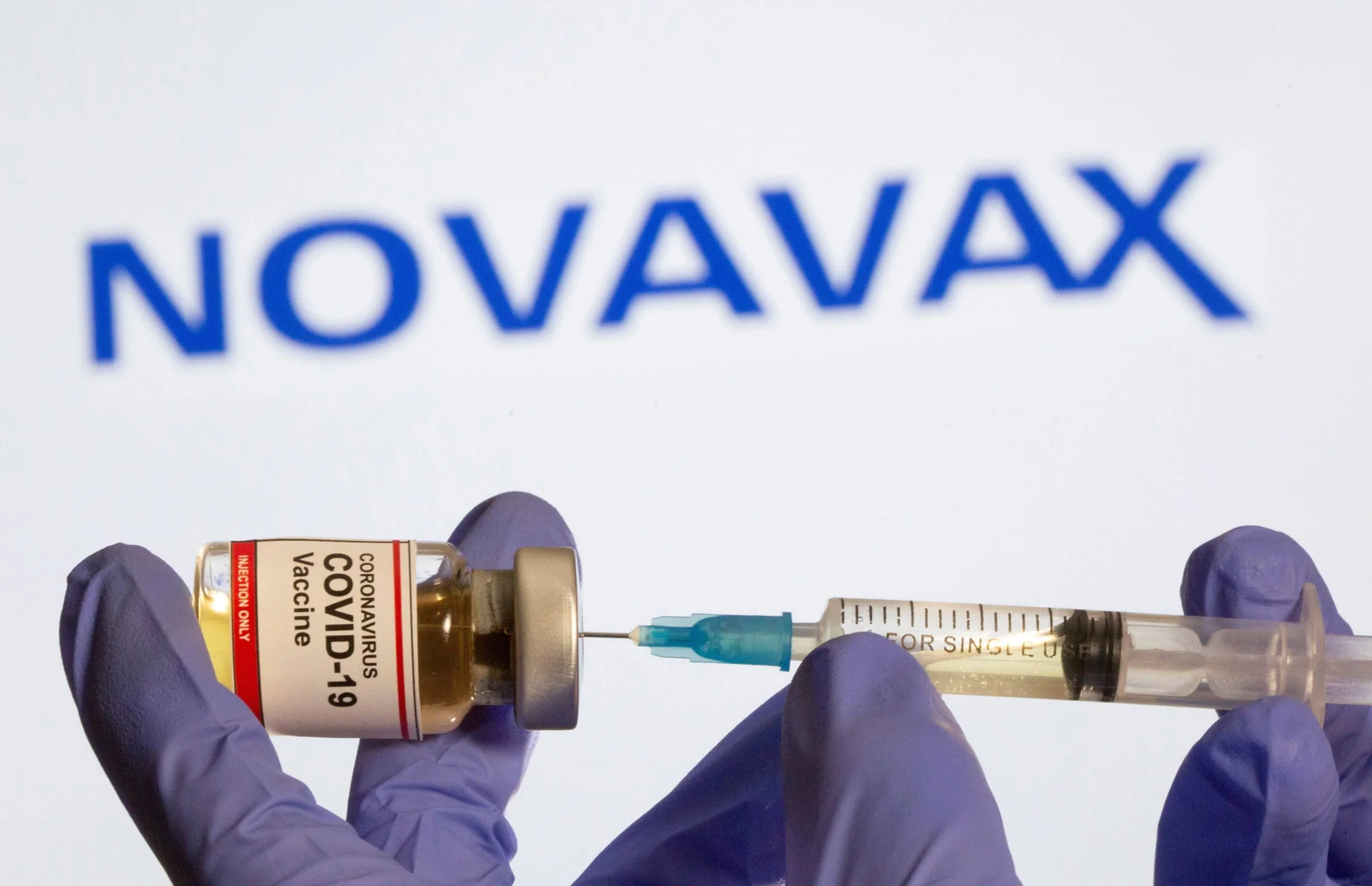 Manos con guantes sostienen un frasco de vacuna contra COVID-19 y una jeringa frente al logo de Novavax.