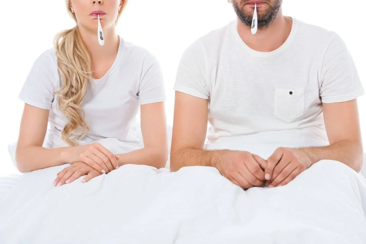 Dos personas sentadas en cama con termómetros en la boca, tomándose la temperatura por enfermedad.