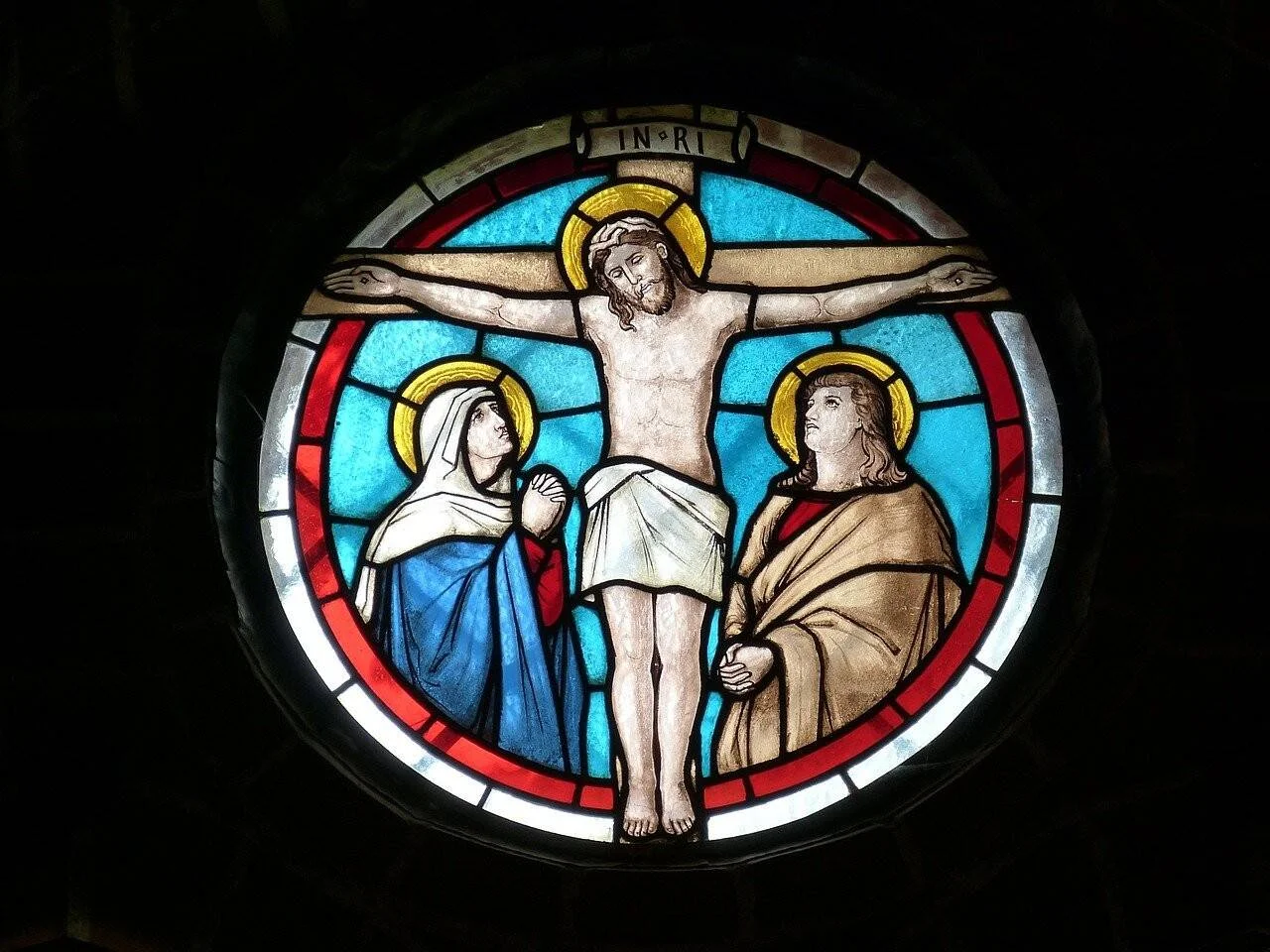 Vitral redondo de iglesia con la crucifixión de Cristo flanqueado por dos figuras con aureolas doradas sobre fondo azul.