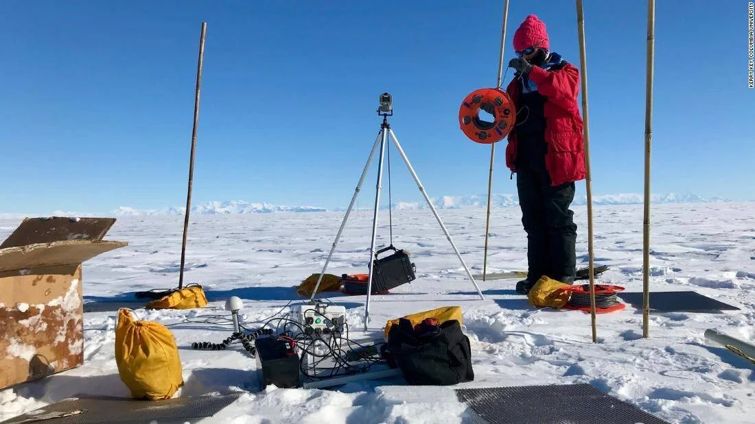Científico equipado con ropa polar sostiene un carrete junto a instrumentos de medición sobre el hielo antártico bajo cielo despejado.