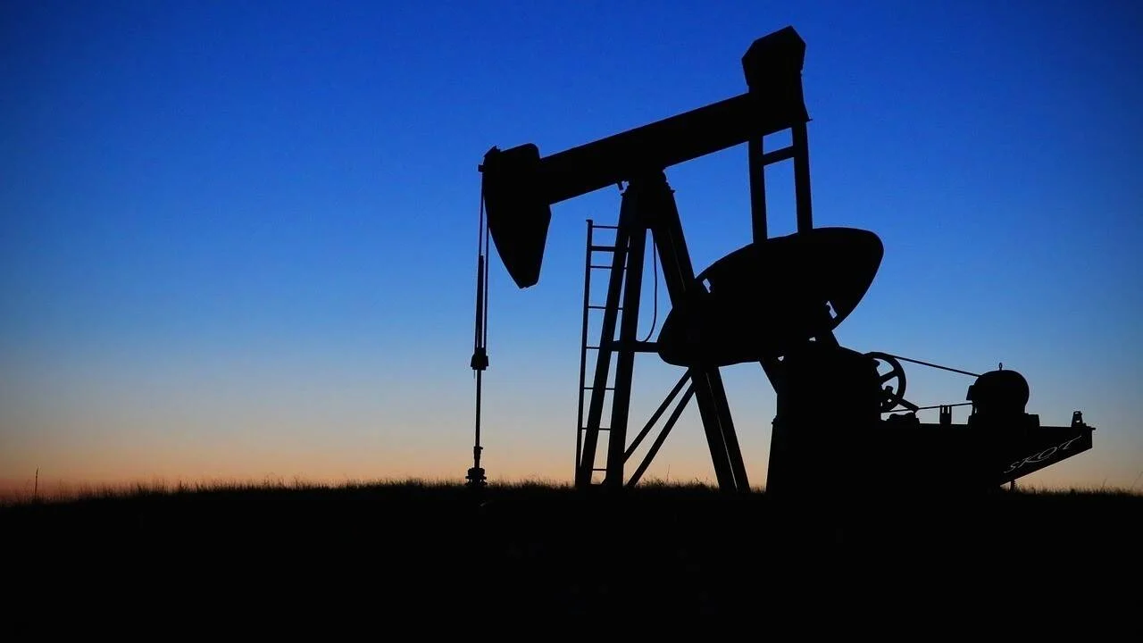 Silueta de una bomba extractora de petróleo en campo abierto, recortada contra un cielo degradado entre azul y naranja al atardecer.
