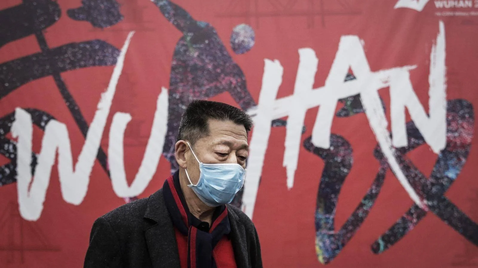 Un hombre con mascarilla quirúrgica camina frente a un gran cartel rojo con la inscripción 'Wuhan'.