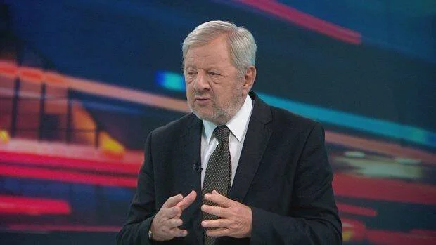 Hombre mayor con traje oscuro y corbata habla gesticulando en un estudio de televisión con fondo iluminado.