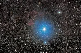 Estrella azul muy luminosa rodeada de un halo celeste y nebulosa rojiza en un campo estelar profundo.