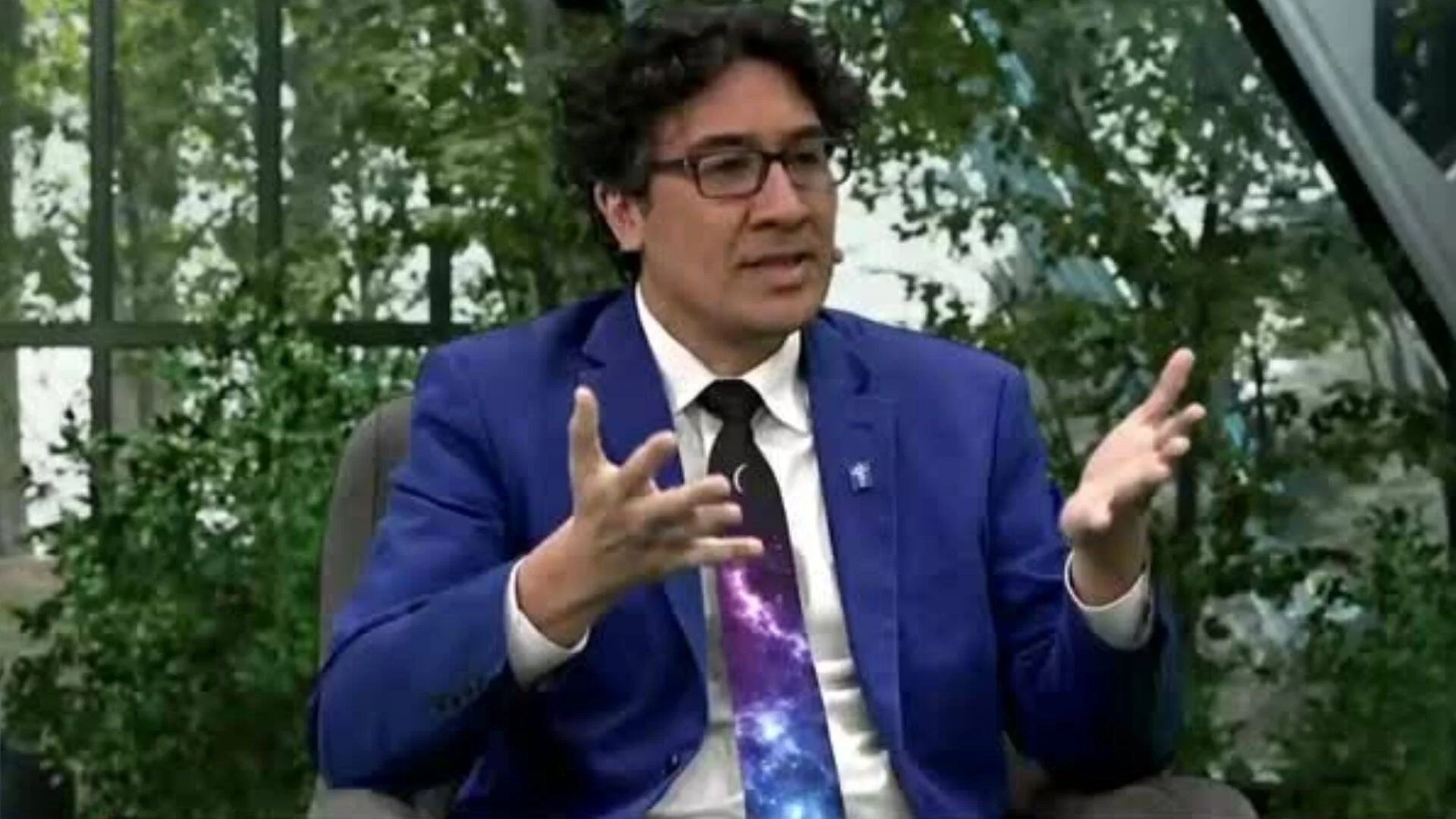 Hombre con anteojos, saco azul y corbata con estampado galáctico gesticula mientras habla en una entrevista televisiva.