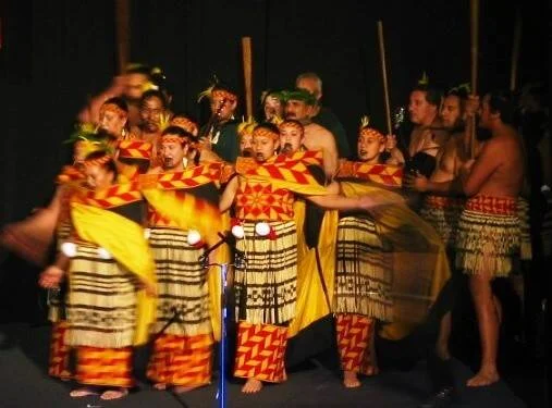 Grupo de bailarines con vestimentas tradicionales de colores rojo, amarillo y naranja realizan una danza cultural en un escenario oscuro.