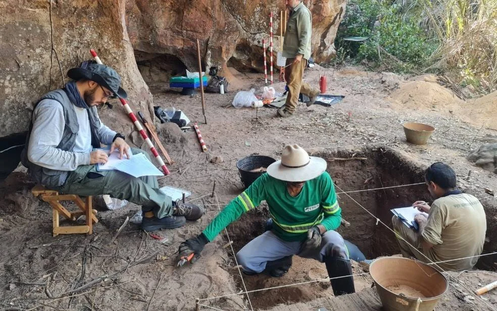 Arqueólogos trabajan en una excavación delimitada con cuerdas junto a una formación rocosa, tomando notas y extrayendo muestras del suelo.