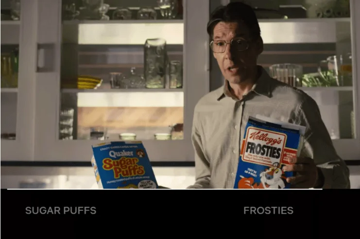Hombre con gafas sostiene una caja de Quaker Sugar Puffs y una bolsa de Kellogg's Frosties frente a una cocina.