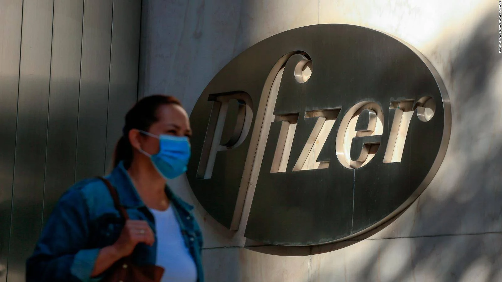 Una mujer con mascarilla azul pasa frente al logo metálico de Pfizer en la fachada de un edificio.