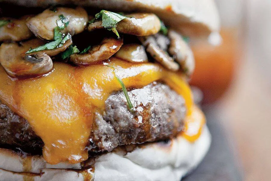 Hamburguesa jugosa con queso cheddar fundido, hongos salteados y hierbas frescas sobre pan artesanal.