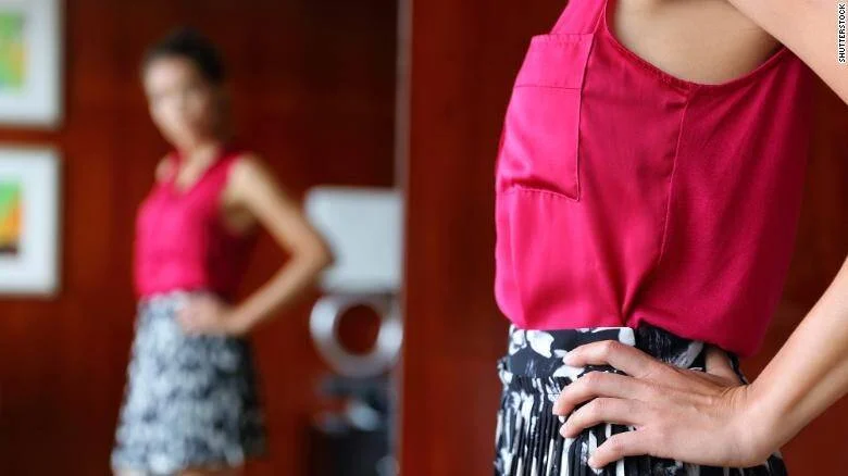 Mujer con blusa rosa y falda estampada se mira al espejo con las manos en la cintura.