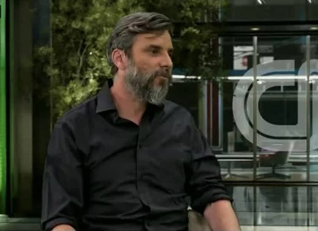 Hombre de mediana edad con barba canosa y camisa negra sentado en un estudio de televisión.