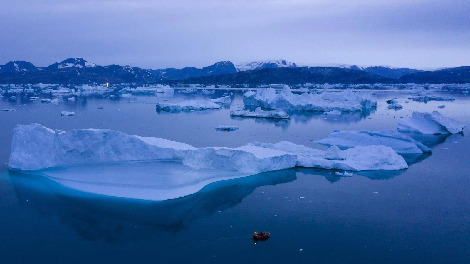 Una pequeña lancha navega entre grandes icebergs flotantes en aguas árticas al atardecer, con montañas nevadas al fondo.