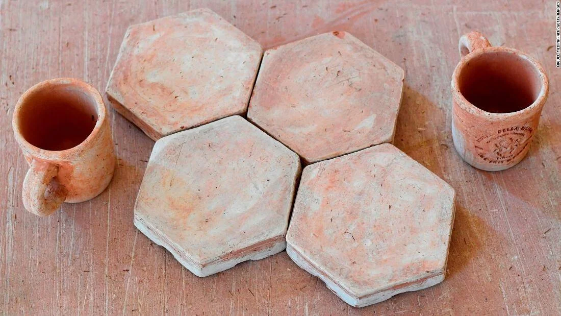 Cuatro baldosas hexagonales de terracota junto a dos pequeñas tazas de barro con líquido oscuro en su interior.
